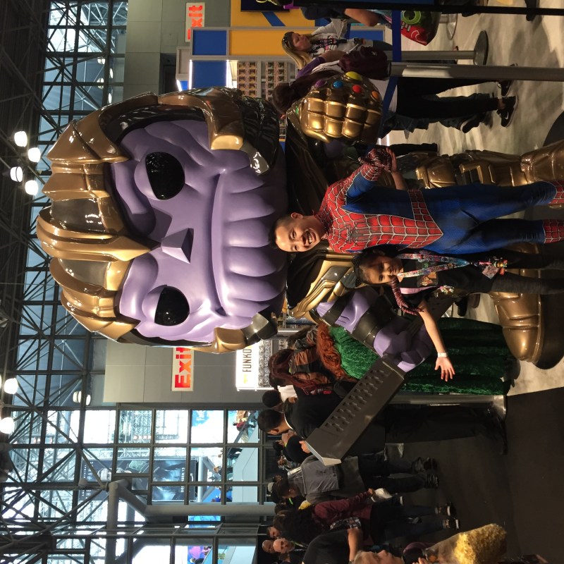 Man wearing a spiderman costume and a girl having a photo opportunity with a doll version of Thanos from The Avengers in an exhibit.