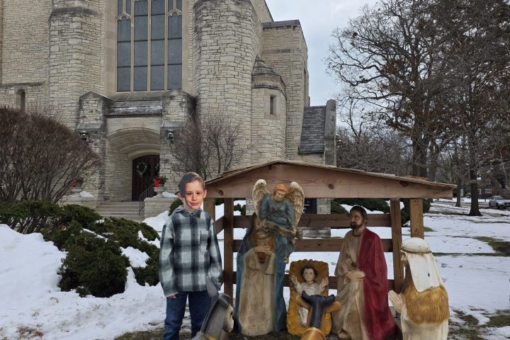 Child standing with nativity figures outside a stone church on a snowy day.