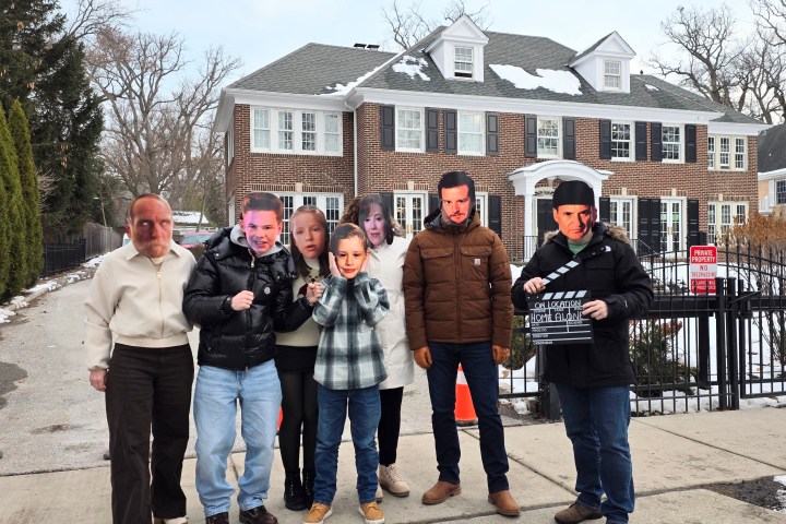 Group of seven people wearing celebrity face masks outside a large house.