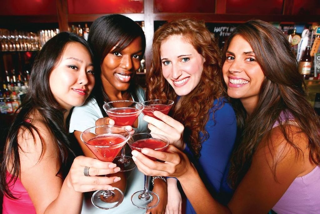 Four women smiling and holding cocktails in a bar setting.